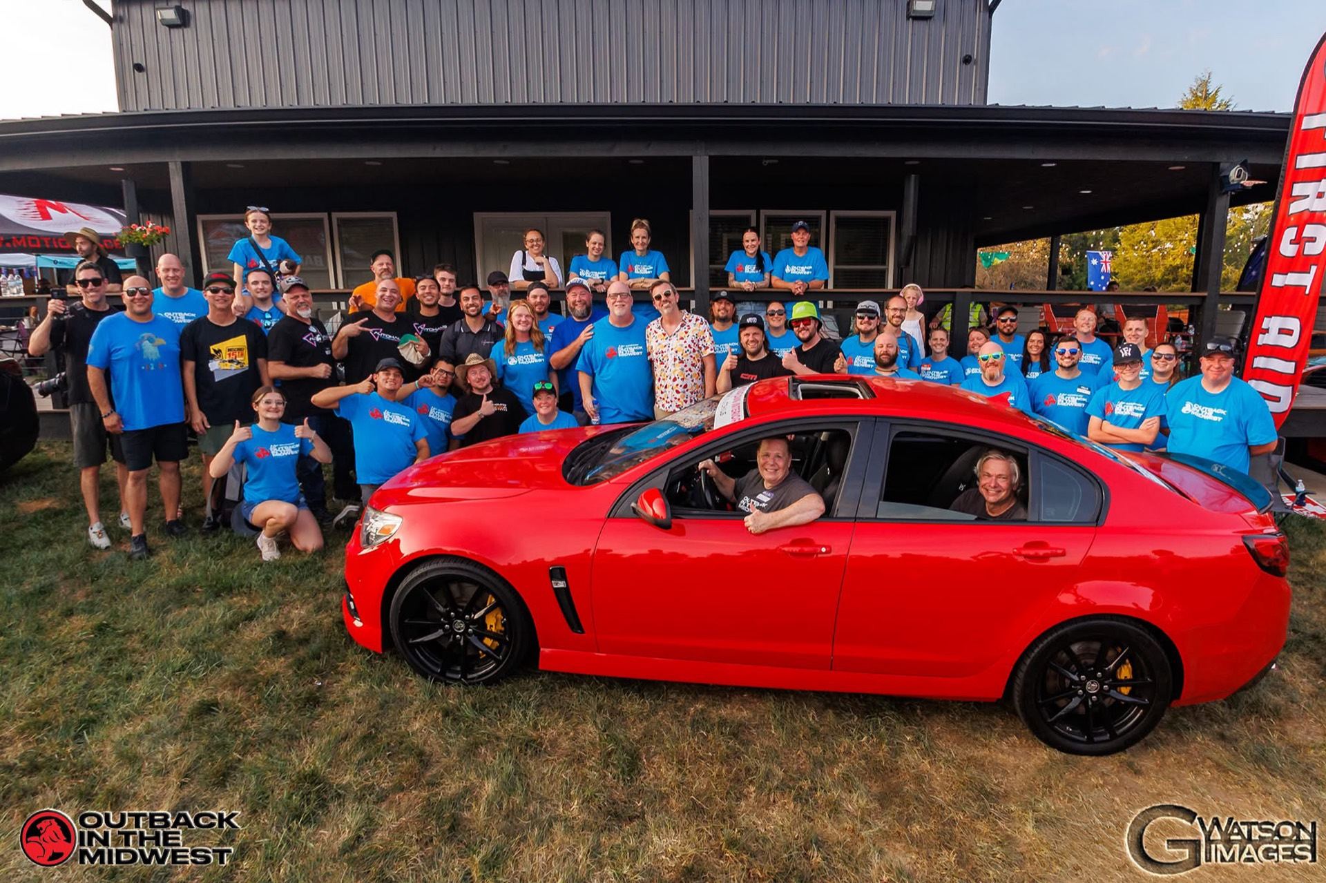 Large group photo with the red HSV in front of the venue, branded for the 10th Anniversary Outback in the Midwest
